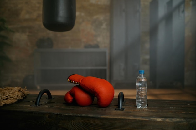 Guantes rojos de boxeo, botella plástica de agua y saco de boxeo en el gimnasio deportivo