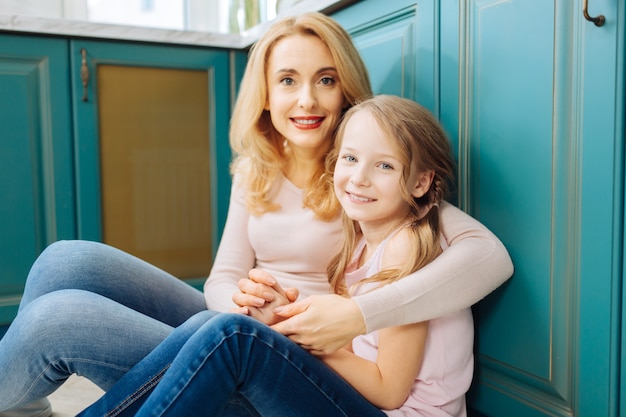 Guapa alegre rubia madre e hija sonriendo y sentada en el suelo de la cocina y abrazándose