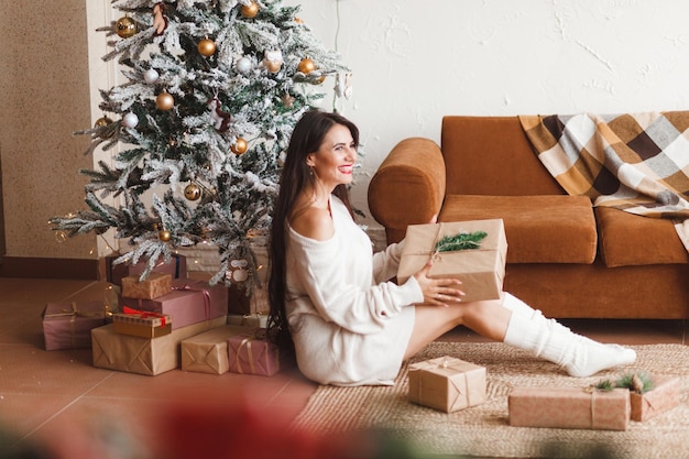 Una guapa morena con un suéter de punto sostiene una caja de regalo en el interior decorado de la casa durante las vacaciones de Navidad y Año Nuevo en el interior