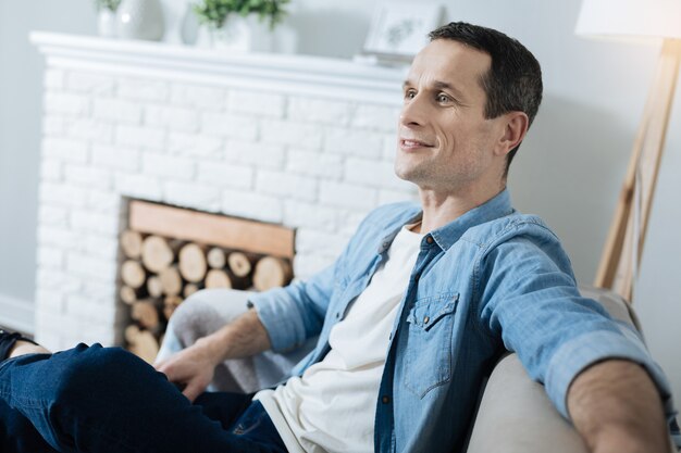 Foto guapo alegre joven de pelo oscuro sonriendo y sentado en el sofá y relajándose