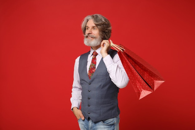 Guapo anciano de pelo gris bigote barbudo hombre en camisa chaleco corbata posando aislado sobre fondo rojo. Concepto de estilo de vida de las personas. Simulacros de espacio de copia. Sosteniendo una bolsa de paquete con compras después de ir de compras.