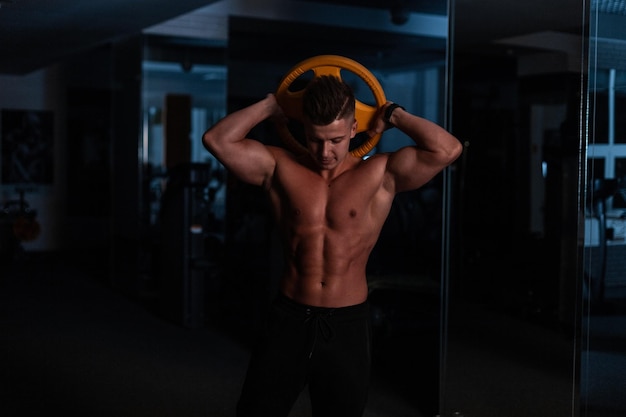 Guapo atleta joven fuerte en un hermoso cuerpo con músculos haciendo ejercicio en el gimnasio en la oscuridad