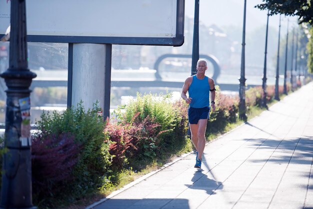 guapo atleta senior trotar y hacer ejercicio matutino con el amanecer y la ciudad en segundo plano