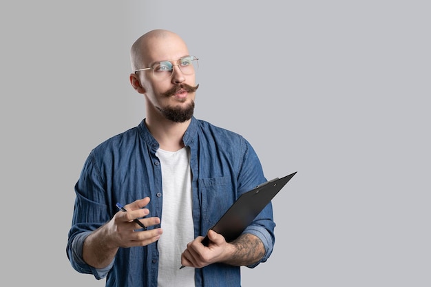 Guapo barba calva con bigote hombre vestido con ropa casual sosteniendo portapapeles aislado sobre gris