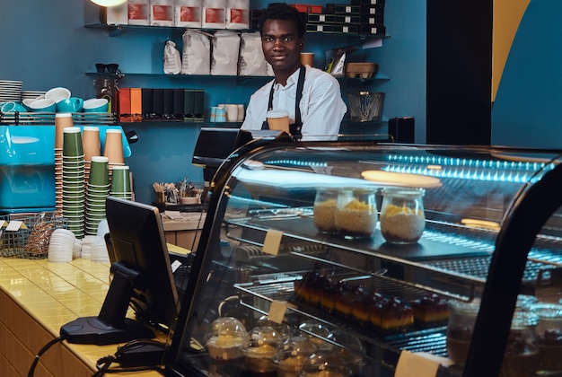 Guapo barista afroamericano en uniforme sostiene una taza de café mientras está de pie en su cafetería.