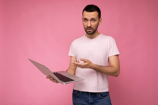 Guapo brunet sorprendido y asombrado que sostiene la computadora portátil que muestra al netbook mirando a la cámara en la camiseta en fondo rosado aislado.