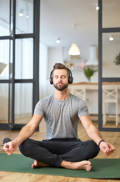 Foto guapo caballero en auriculares inalámbricos disfrutando de música relajante y haciendo ejercicio de meditación mientras está sentado en una estera de yoga en casa