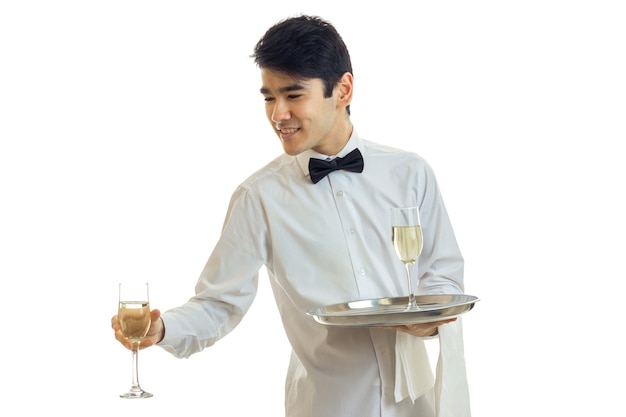 Guapo camarero con una camisa blanca y con una mariposa se extiende copa de vino aislado en la pared blanca