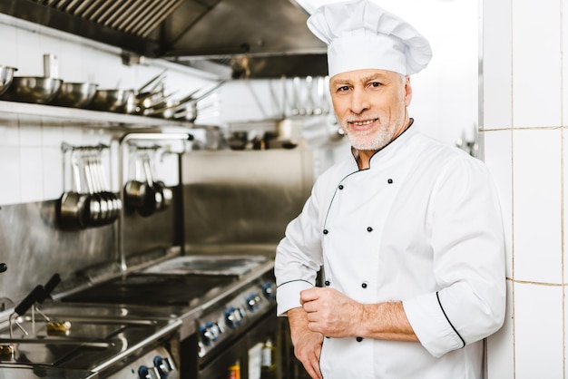 guapo chef masculino sonriente en uniforme y gorra mirando a la cámara en la cocina del restaurante con copia