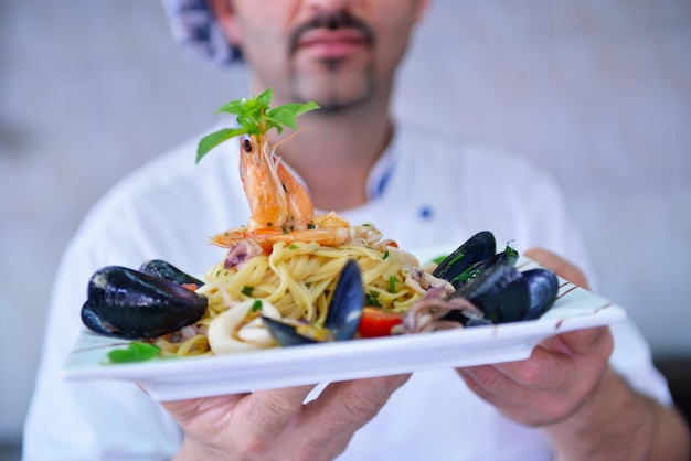 Guapo chef vestido con uniforme blanco decorando ensalada de pasta y pescado de marisco en la cocina moderna