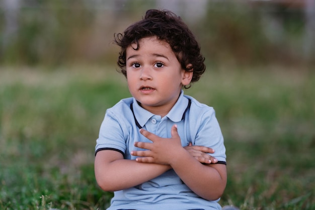 Guapo chico español sentado en la hierba se abraza a sí mismo Lindo niño hispano al atardecer disfruta del verano al aire libre Infancia y estilo de vida de los niños