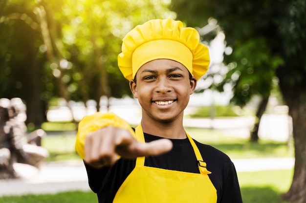 Guapo cocinero adolescente africano le señala a usted Cocinero de niño negro con sombrero de chef y uniforme de delantal amarillo sonriendo y señalándole al aire libre Publicidad creativa para café o restaurante