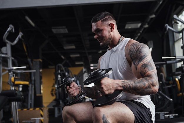 Guapo culturista musculoso tatuado sosteniendo pesas pesadas sentado en el gimnasio