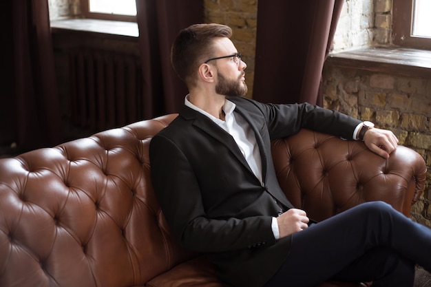 Guapo y elegante hombre de negocios barbudo confiado en un traje inteligente y gafas sentado en un sofá en la oficina