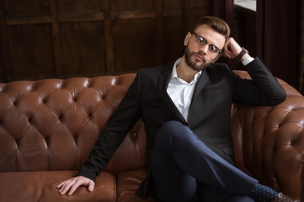 Foto guapo y elegante hombre de negocios barbudo confiado en un traje inteligente y gafas sentado en un sofá en la oficina