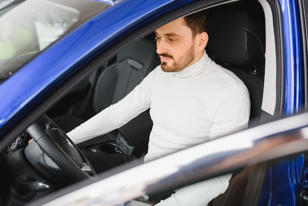 Guapo empresario barbudo está sentado en un coche nuevo en concesionario de coches