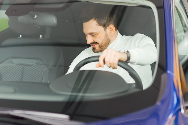 Guapo empresario barbudo está sentado en un coche nuevo en concesionario de coches