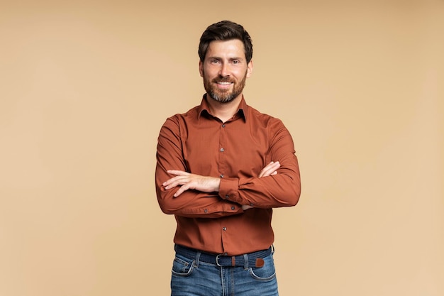 Guapo empresario de mediana edad sonriente posando con los brazos cruzados aislado sobre un fondo beige