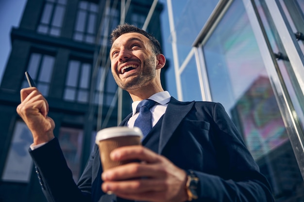 Guapo empresario morena mostrando emociones de felicidad mientras pasa tiempo al aire libre con café y teléfono inteligente