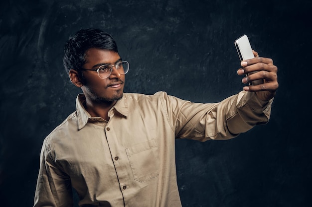 Guapo estudiante hindú sonríe y hace selfie en teléfono en estudio sobre fondo negro