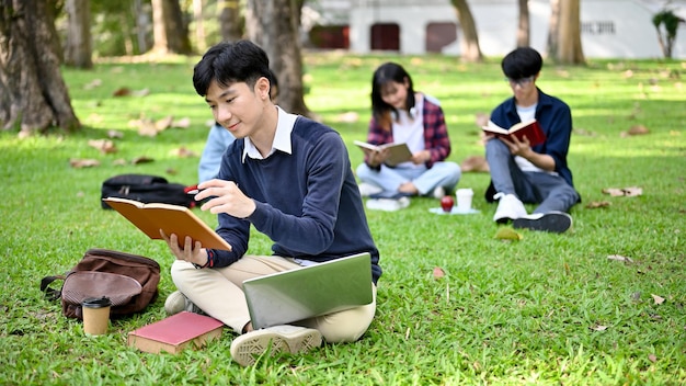 Guapo estudiante universitario masculino asiático leyendo un libro y haciendo la tarea en el parque del campus