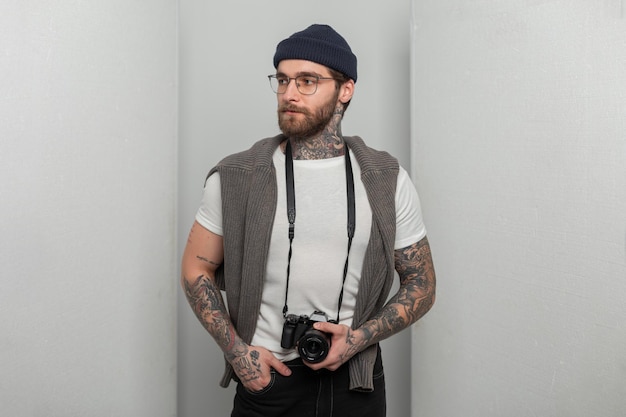 Foto guapo exitoso fotógrafo profesional hipster con gafas con un tatuaje en un sombrero de punto con un suéter sostiene una cámara fotográfica y se encuentra en el estudio