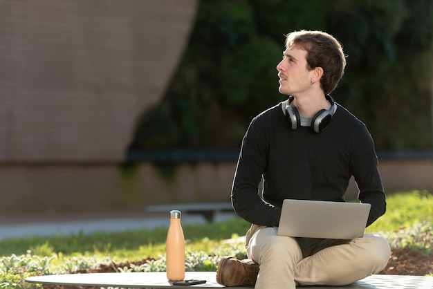 Guapo freelancer trabajando en su computadora portátil al aire libre, sentado en un banco en el parque con hermosa luz del sol