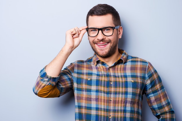 Guapo con gafas. Apuesto joven ajustando gafas y sonriendo mientras está de pie contra el fondo gris