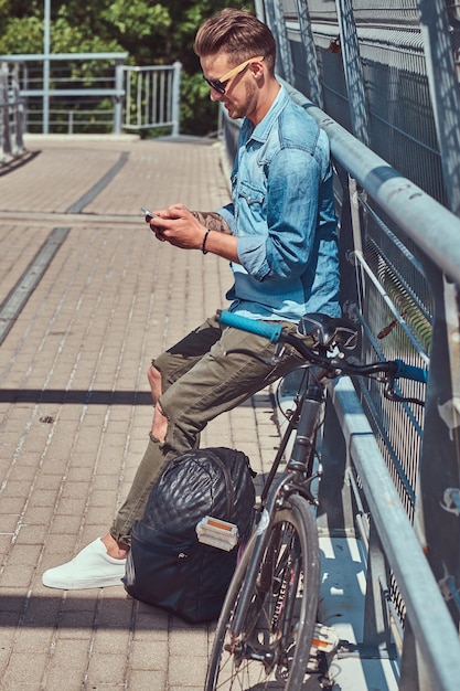 Guapo hipster con un elegante corte de pelo con gafas de sol descansando después de andar en bicicleta, usando un teléfono inteligente.