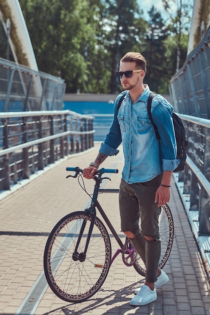 Guapo hipster de moda con un elegante corte de pelo en gafas de sol caminando con bicicleta al aire libre.