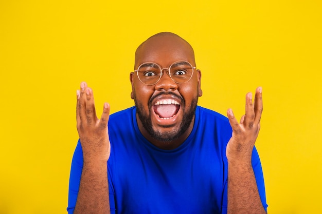 Guapo hombre afro brasileño con gafas camisa azul sobre fondo amarillo Primer plano cerrado photoxACelebrating sorprendido sorprendido wow éxito alegría manos abiertas asombrado gritando de alegría