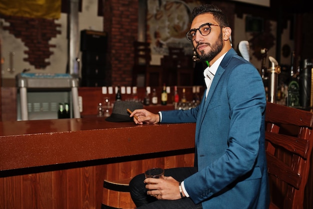 Guapo hombre árabe bien vestido con vaso de whisky y cigarro posado en el pub