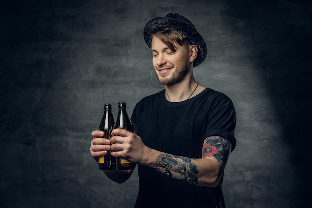 Guapo hombre barbudo con brazos tatuados, vestido con una camiseta negra y sombrero de copa sostiene cerveza artesanal embotellada.