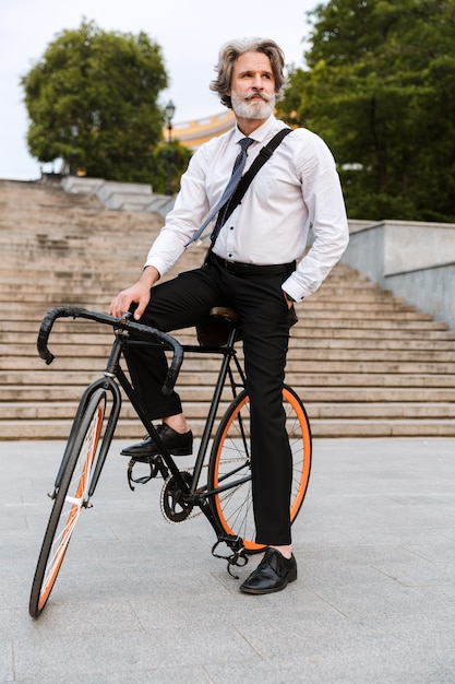 Guapo hombre barbudo confiado en traje, llevando maletín caminando al aire libre con bicicleta