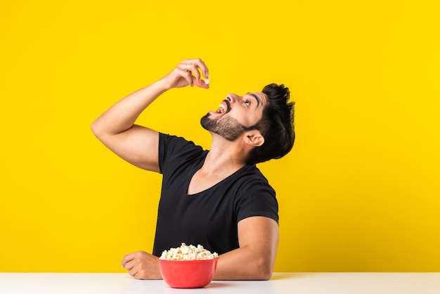 Guapo hombre barbudo Indin comiendo palomitas de tazón rojo mientras está sentado en la mesa contra el fondo amarillo