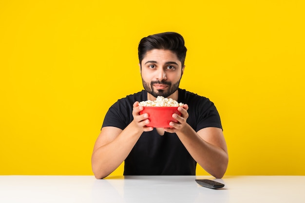 Guapo hombre barbudo Indin comiendo palomitas de tazón rojo mientras está sentado en la mesa contra el fondo amarillo