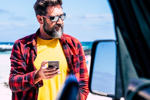 Guapo hombre caucásico con barba usa un teléfono moderno fuera del coche con olas de playa y mar en escena