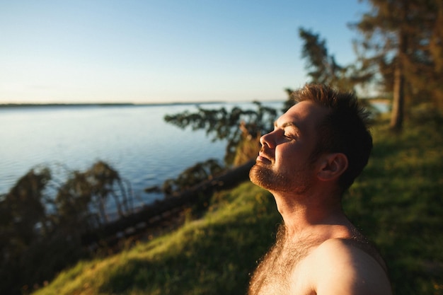 Foto guapo hombre caucásico barbudo con los ojos cerrados de pie sobre un acantilado en la hermosa puesta de sol en segundo plano.