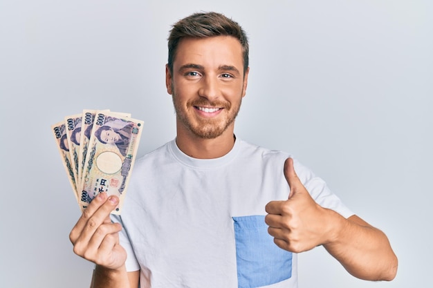 Guapo hombre caucásico sosteniendo billetes de 5000 yenes japoneses sonriendo feliz y positivo pulgar hacia arriba haciendo excelente y signo de aprobación