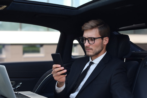 Guapo hombre de confianza en traje completo mirando su teléfono inteligente mientras está sentado en el coche y usando la computadora portátil.