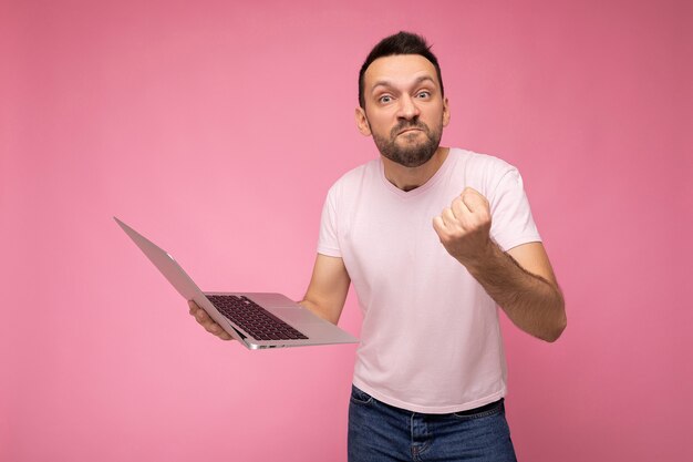 Guapo hombre enojado y furioso con ordenador portátil mostrando puño mirando a cámara en camiseta sobre fondo rosa aislado.