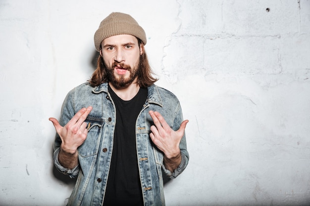 Guapo hombre hipster barbudo con sombrero vestido con chaqueta de jeans posando sobre la pared mientras gesticula con las manos