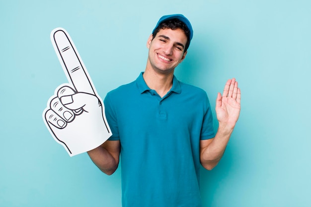 Guapo hombre hispano sonriendo felizmente agitando la mano dándole la bienvenida y saludando el concepto de fanático número uno