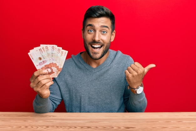 Guapo hombre hispano sosteniendo pesos colombianos apuntando con el pulgar hacia el lado sonriendo feliz con la boca abierta