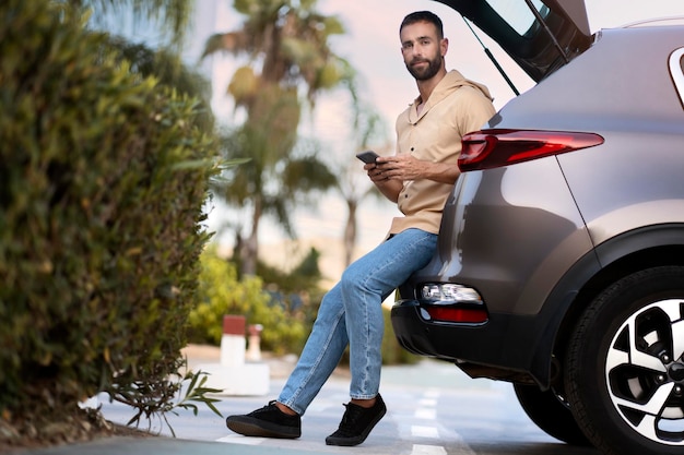 Guapo hombre latino pensativo usando teléfono móvil mirando hacia otro lado planeando un viaje por carretera sentado en el baúl del auto