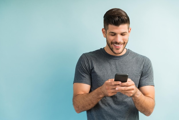 Guapo hombre latino sonriendo mientras envía mensajes de texto en el teléfono inteligente contra un fondo liso