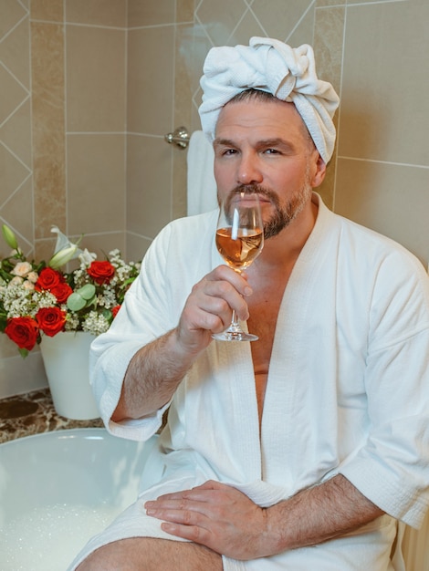 Guapo hombre maduro gay en bata de baño blanca y una toalla en la cabeza sentado en el baño con espuma