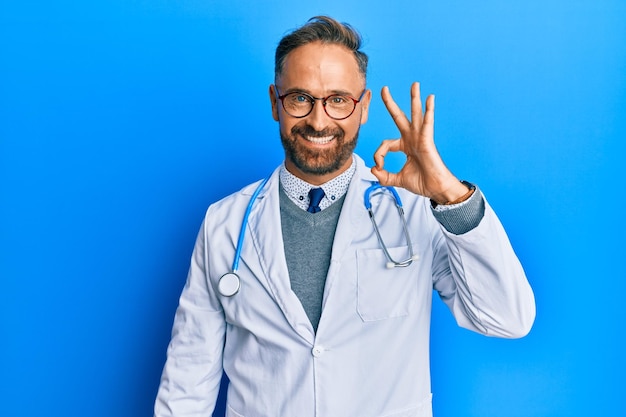 Guapo hombre de mediana edad con uniforme médico y estetoscopio sonriendo positivamente haciendo bien el signo con la mano y los dedos. expresión exitosa.