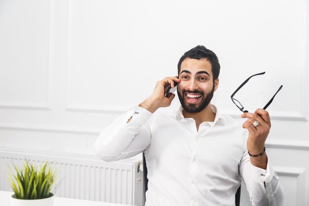 Guapo hombre de negocios feliz barbudo usa camisa blanca sostiene anteojos en la mano habla por teléfono en el interior de la oficina blanca