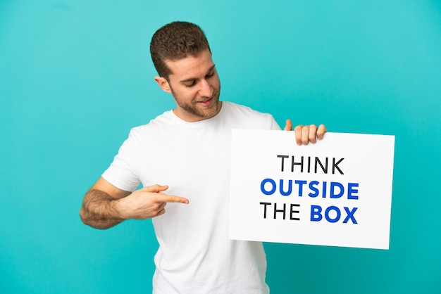 Guapo hombre rubio sobre fondo azul aislado sosteniendo un cartel con el texto Piense fuera de la caja y apuntando